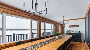 Une longue table à manger en bois, entourée de chaises, de grandes fenêtres offrant une vue sur un paysage enneigé, un lustre et une enseigne indiquant « CHAMBRES D’HÔTES ET PETIT-DÉJEUNER ».