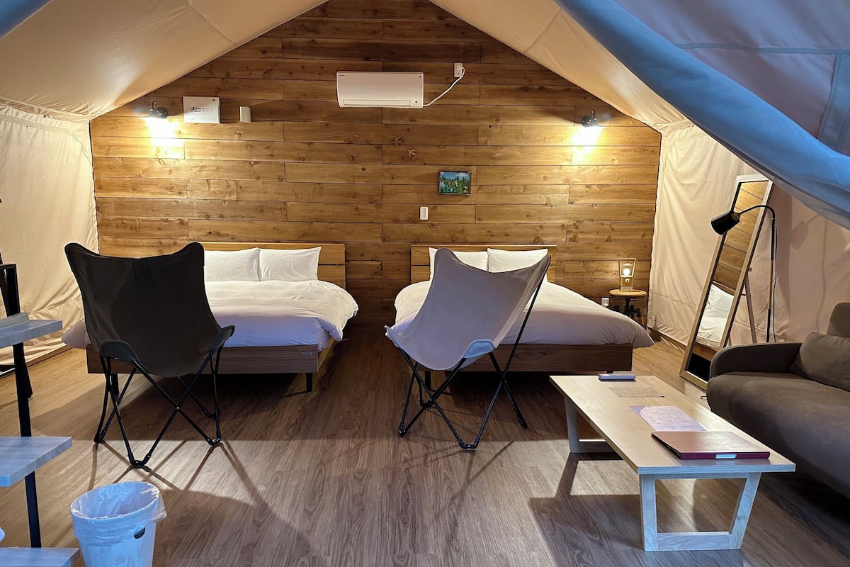 A cozy tented accommodation with two beds, foldable chairs, a small table, and a lamp.