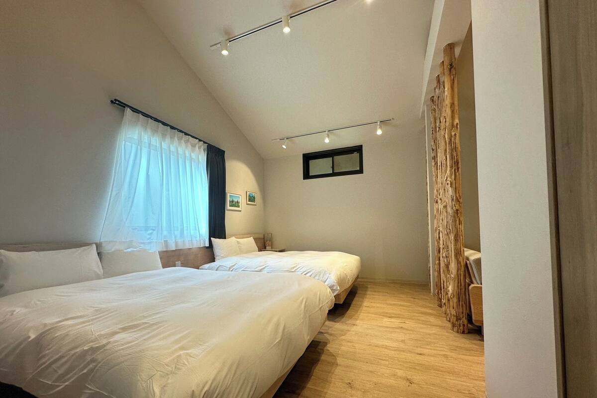 A bedroom with two beds, a window with sheer curtains, a small wall-mounted picture, and a wooden floor.