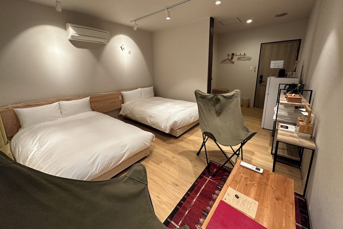 A hotel room with two beds, a desk, a chair, an air conditioner, and a refrigerator.