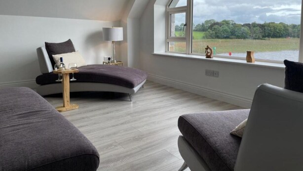 Ein Zimmer mit einer gebogenen Couch, einem kleinen Tisch mit einer Lampe und Seeblick durch ein großes Fenster.