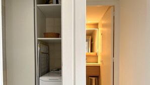 Un couloir étroit avec un panier à linge sur une étagère, un radiateur et un lave-linge blanc.