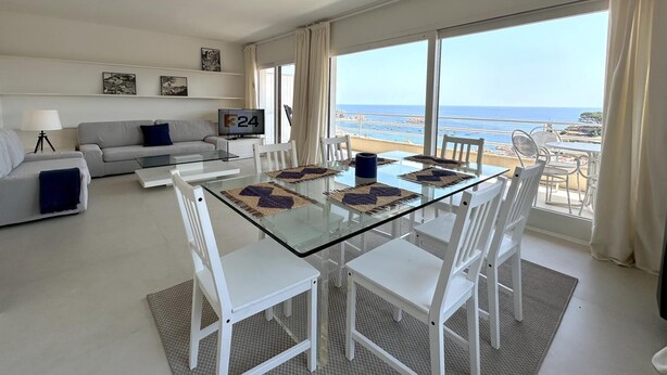 Un salon moderne avec une table à manger au plateau en verre, des chaises blanches, un canapé gris et un téléviseur. De grandes fenêtres offrent une vue sur la plage.