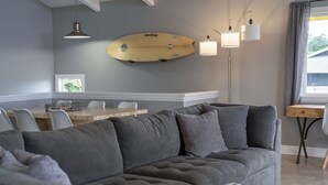 A modern living room with a grey sofa, a wooden dining table with chairs, a surfboard on the wall, and a floor lamp. - Beautiful conveniently located 2-bedroom ocean view home in Kailua-Kona.  (Kailua-Kona)
