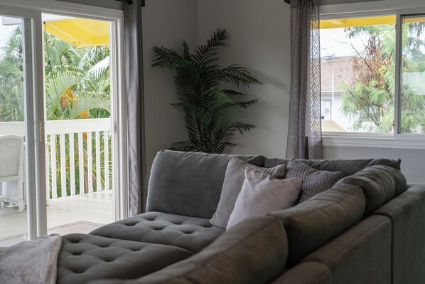 A living room with a grey sofa, a large potted plant, and a view of a balcony with a white railing and palm trees.