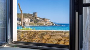 Vista costera desde una ventana, con un muro de piedra, un árbol alto y un edificio con una torre en una colina.
