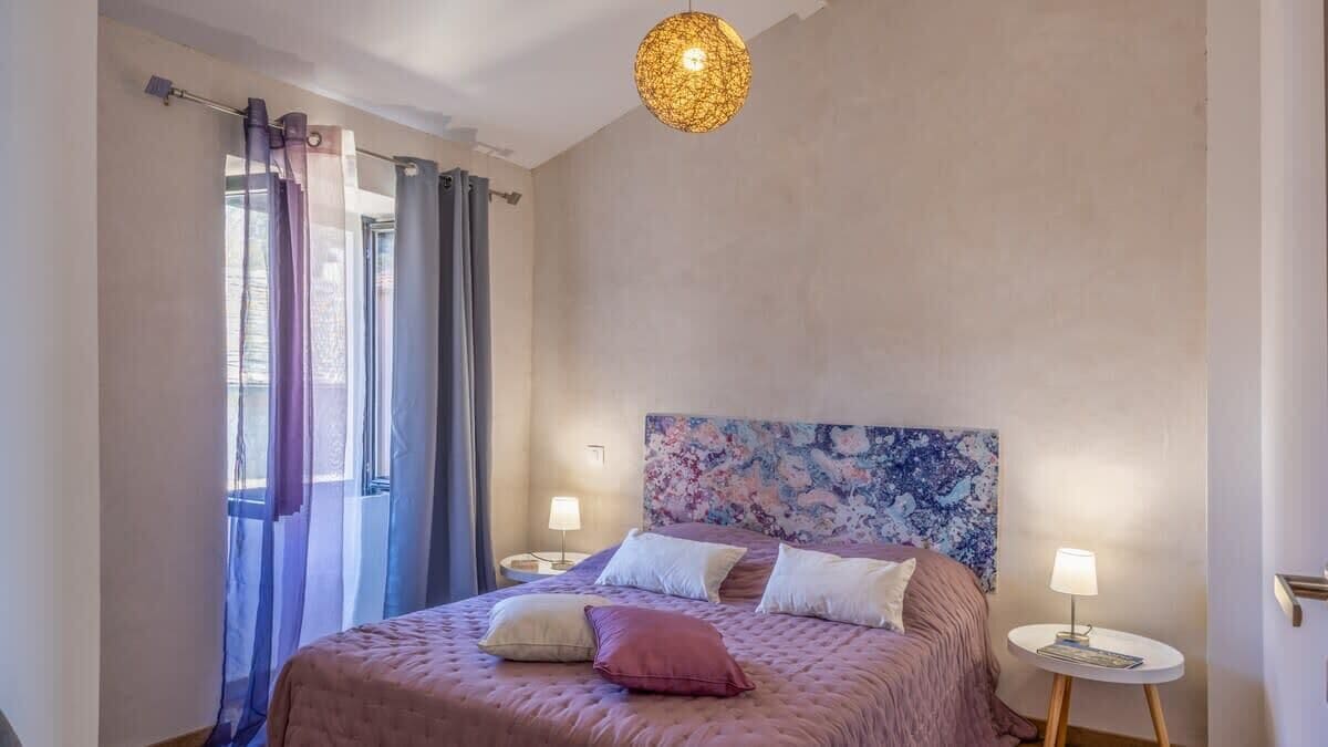 A bedroom with a bed, curtains, a headboard with a floral pattern, bedside lamps, and a hanging light fixture.