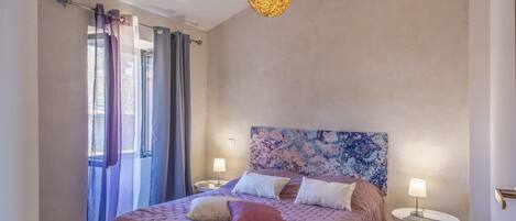 A bedroom with a bed, curtains, a headboard with a floral pattern, bedside lamps, and a hanging light fixture.