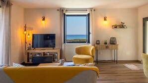 Habitación de hotel con un amplio ventanal que ofrece vistas al mar, un televisor, un sillón amarillo y una pequeña mesa con adornos.