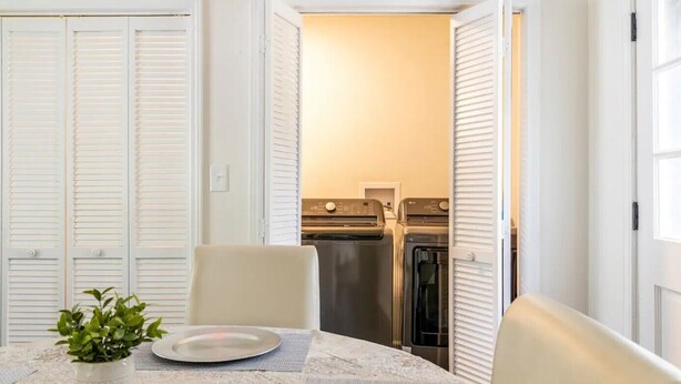 Un comedor con una mesa preparada para dos personas, una planta en maceta y vistas a una cocina con lavadora y horno.