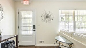 Un interior moderno con una puerta blanca, una pieza decorativa en la pared, una mesa de vidrio y un sofá blanco junto a una ventana.