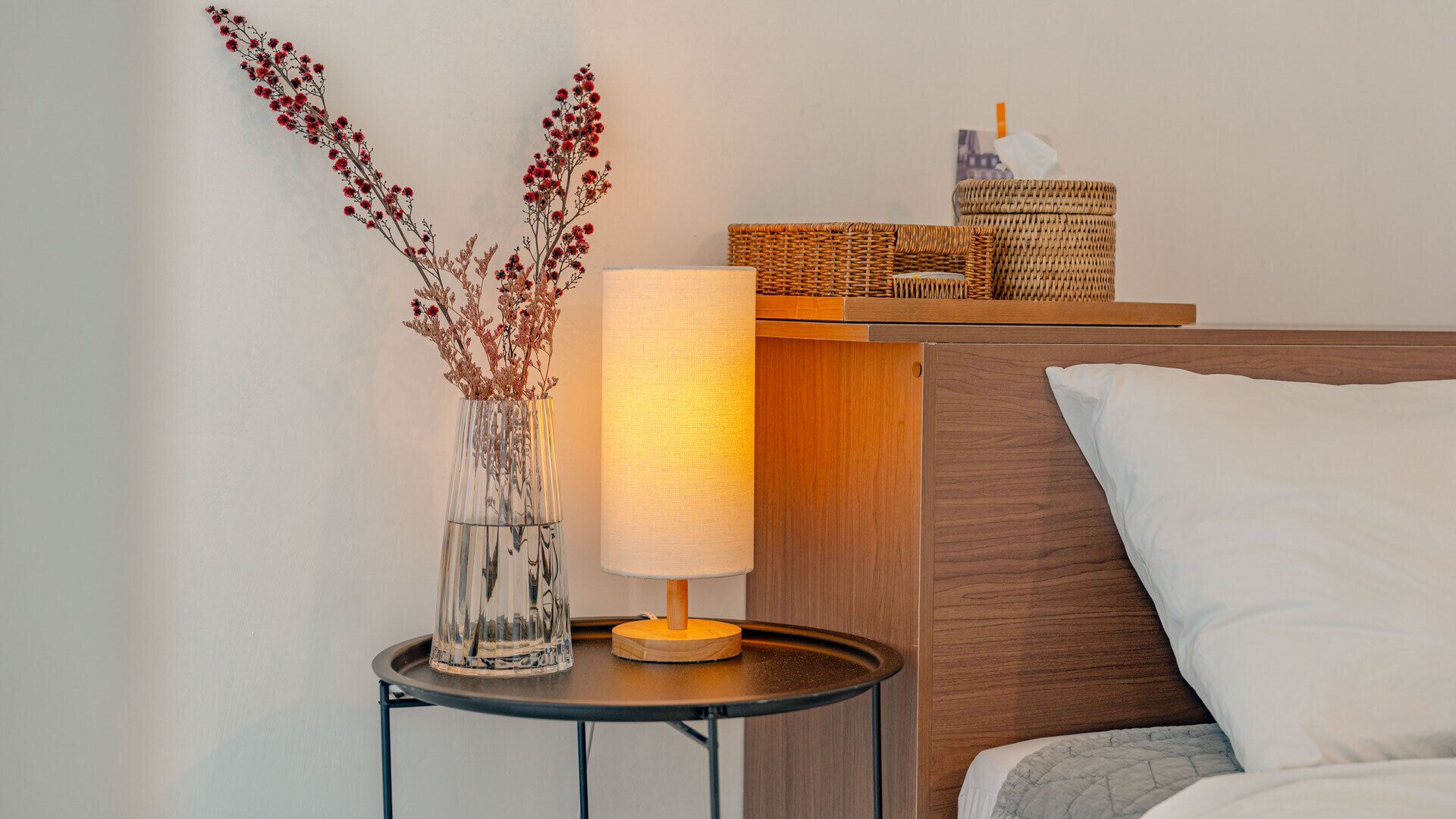 Ein Bett mit weißem Kopfkissen, ein Nachttisch mit Lampe, eine Vase mit Trockenblumen und ein Korbwäscher auf dem Beistelltisch.