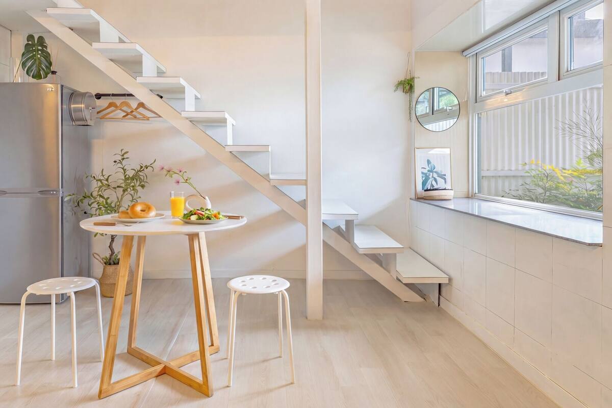 A modern kitchen with a white staircase, a dining table with stools, a refrigerator, and a window with a view.