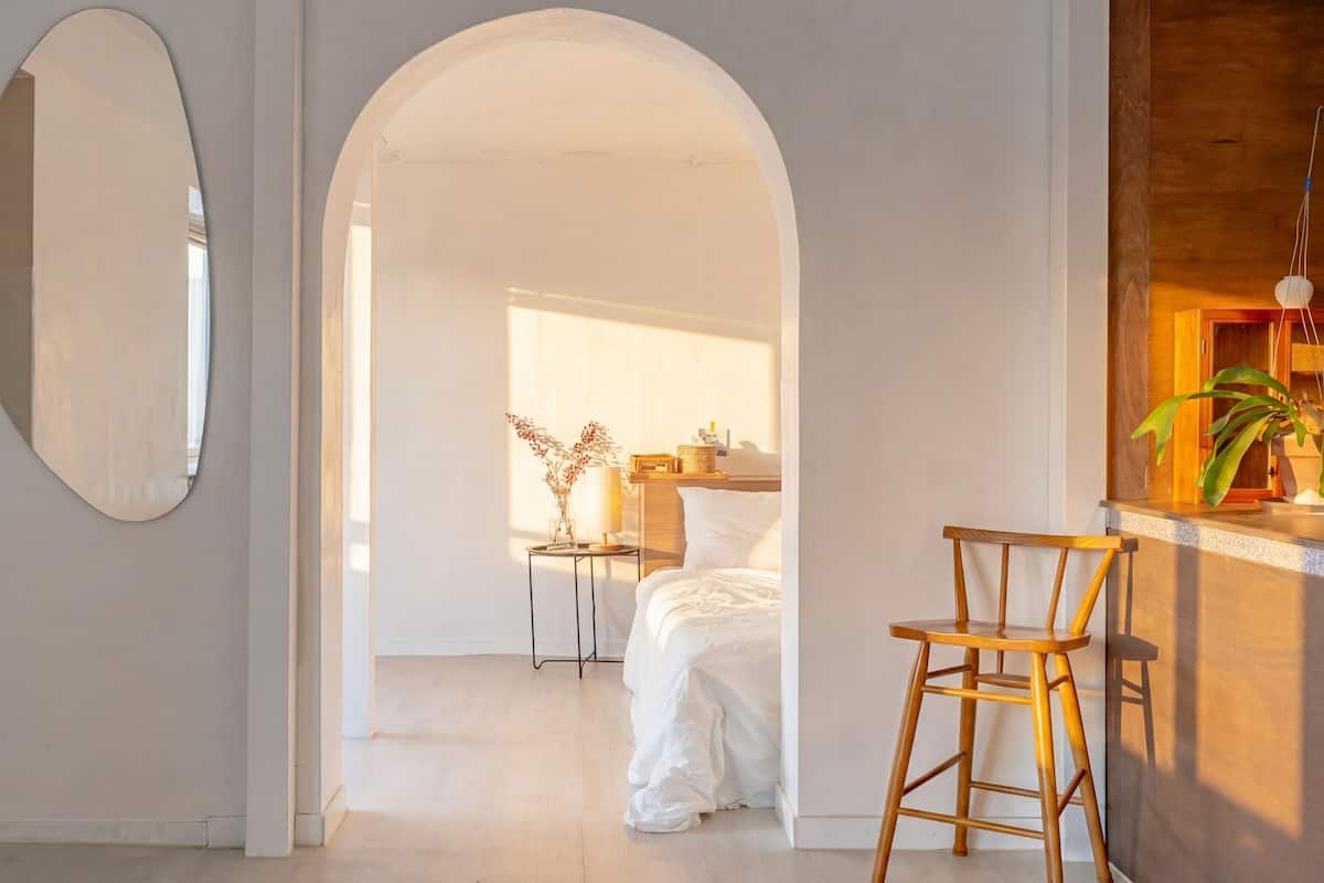 A hotel room with a bed, a wooden chair, a mirror, and a small table with a lamp.