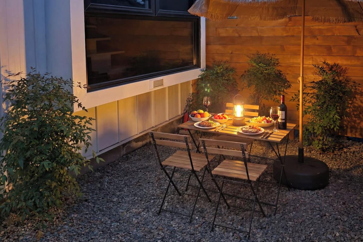 An outdoor dining area with a table set for two, surrounded by potted plants and a thatched roof structure.