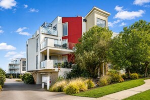 A multi-story residential building with balconies, a red accent wall, and a well-maintained lawn.