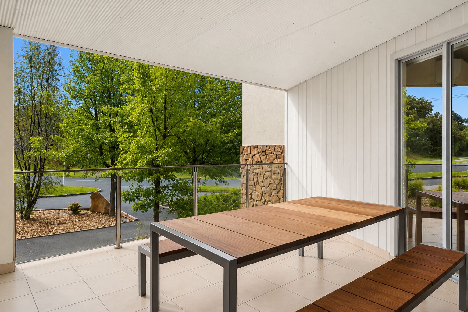 A balcony with a wooden table and benches, a stone wall, and a view of trees and a road.