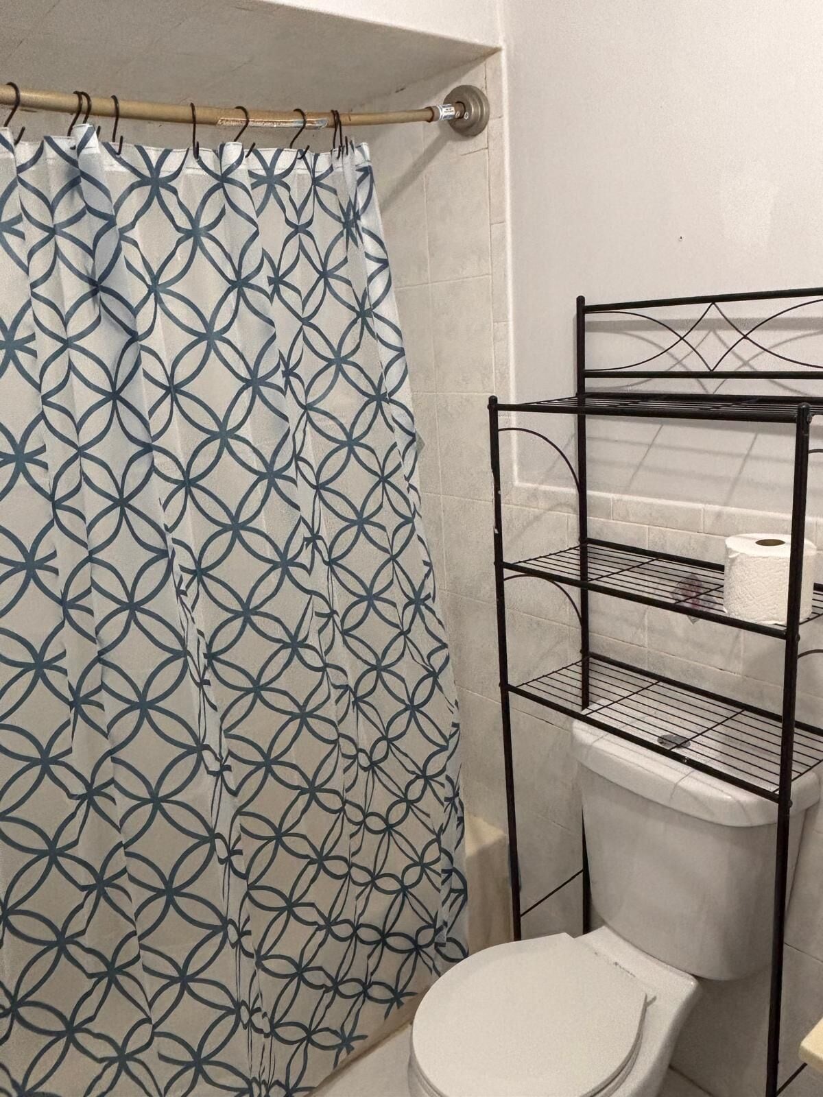 A bathroom with a white toilet, a metal shelf unit with toilet paper, and a patterned shower curtain.