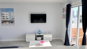 Ein modernes Wohnzimmer mit einem an der Wand befestigten Flachbildfernseher, einer weißen Unterhaltungsanlage, einem Essbereich mit weißen Stühlen und einem großen Fenster mit Vorhängen.