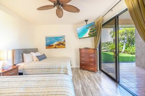 Un dormitorio con dos camas, un ventilador de techo, un televisor, un aparador y una puerta corrediza de vidrio a un jardín.