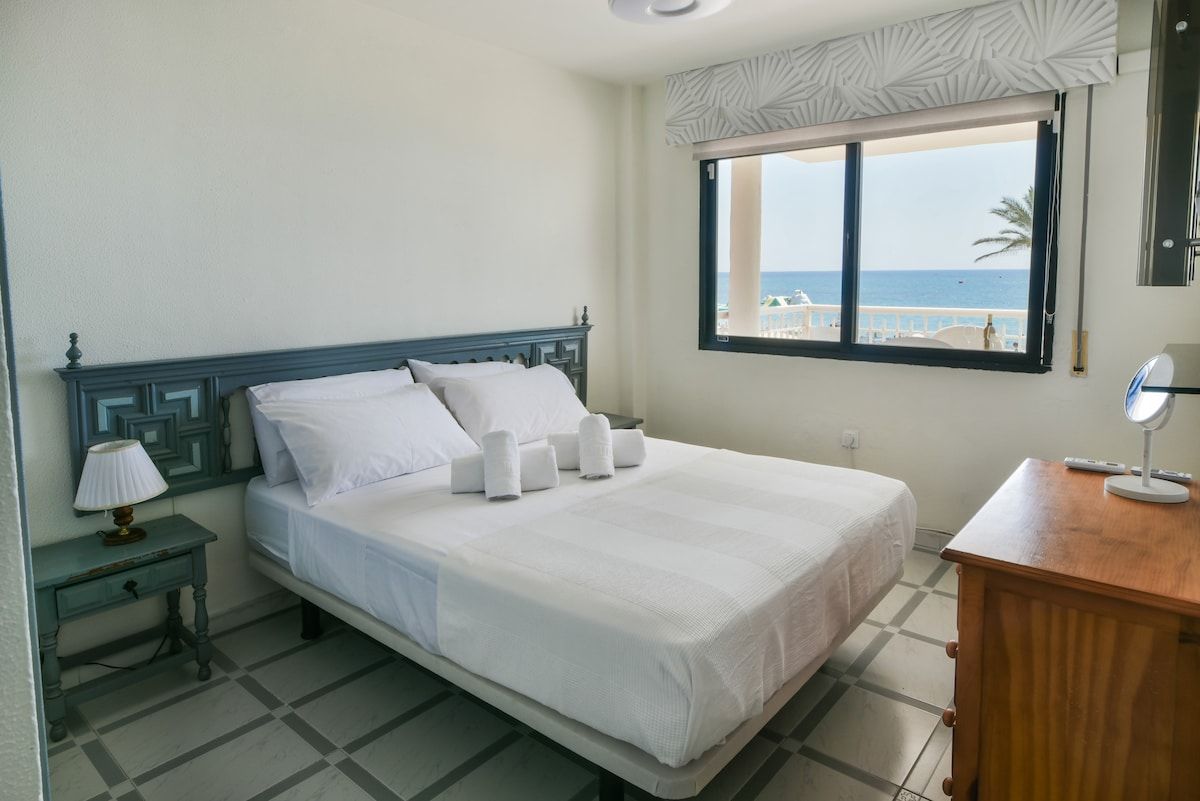 Een hotelkamer met een bed, een nachtkastje met lamp, een houten dressoir en een raam met uitzicht op het strand.