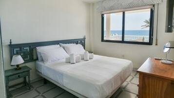 Een hotelkamer met een bed, een nachtkastje met lamp, een houten dressoir en een raam met uitzicht op het strand.