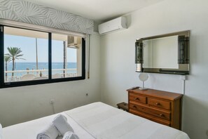 A hotel room with a bed, a wooden dresser, a mirror, and a window with a view of the beach and palm trees. - Benalmadena beachfront location & views (Benalmadena)