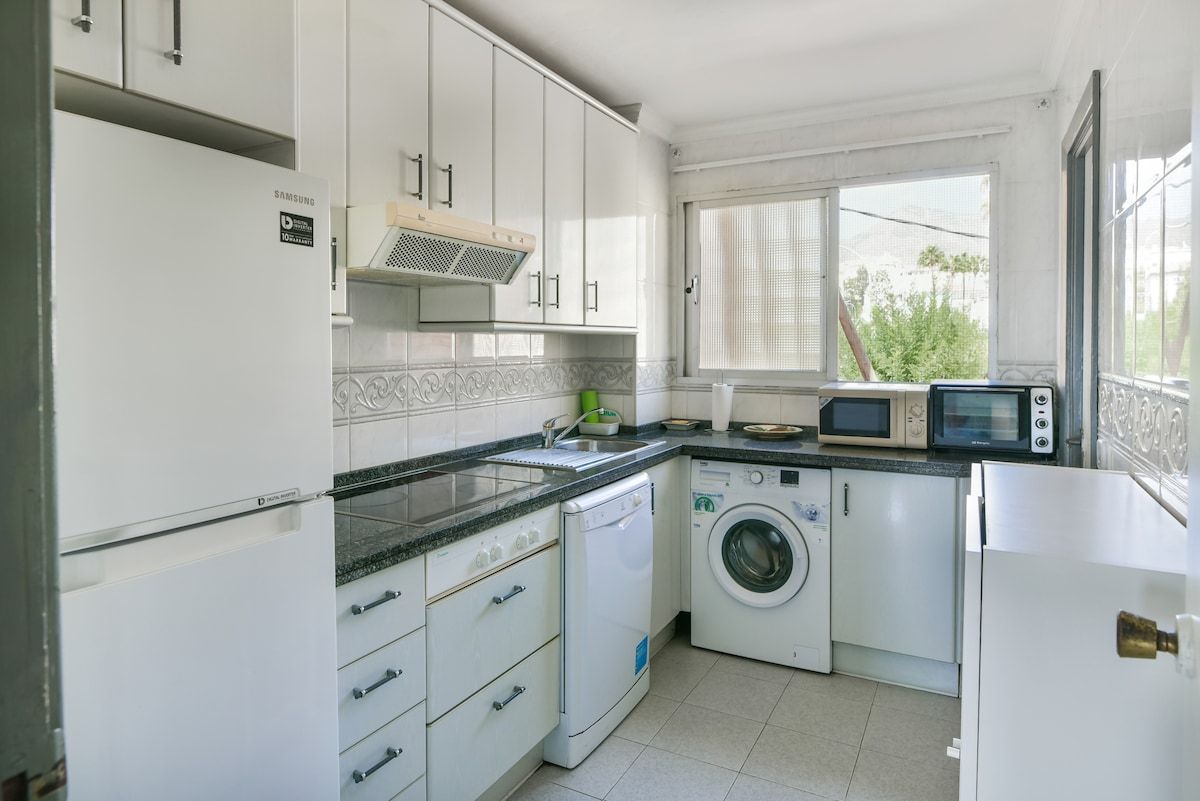 Een kleine keuken met witte kastjes, een wasmachine, een magnetron en een koelkast.