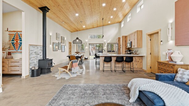 A spacious living area with a large wooden ceiling, a central fireplace, a kitchen with wooden cabinets, and a cozy seating area with a blue sofa.