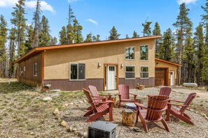 A two-story cabin with a brown exterior, a front door, and windows. There are red Adirondack chairs and a small table in the front yard.