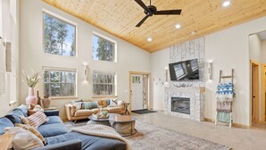A spacious living room with a fireplace, large windows, and a ceiling fan.