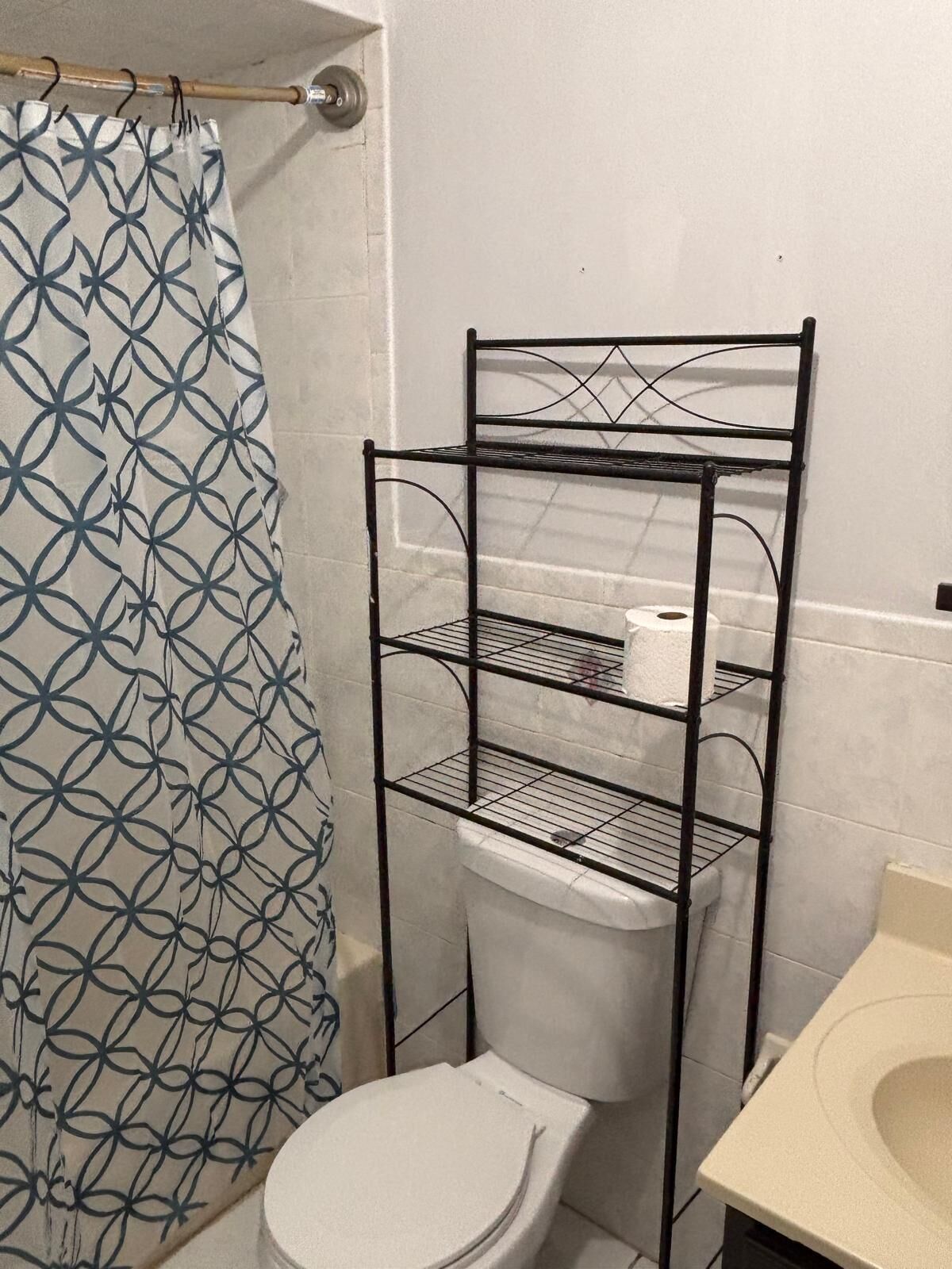 A bathroom with a white toilet, a metal shelf unit, and a patterned shower curtain.