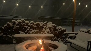 Ein verschneiter Außenbereich mit einem Feuerkorb in der Mitte, umgeben von Schneewällen und Lichterketten.