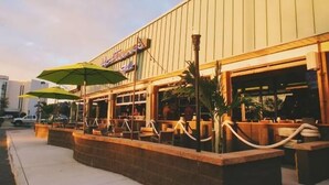 An outdoor seating area with tables and chairs, umbrellas, and a building with a sign. - Sojourn's Sea Views with Balcony Sleeps 4 (Virginia Beach)