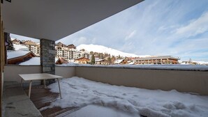 Un balcón con un pilar de piedra, una mesa de madera y vistas a una montaña nevada y edificios.