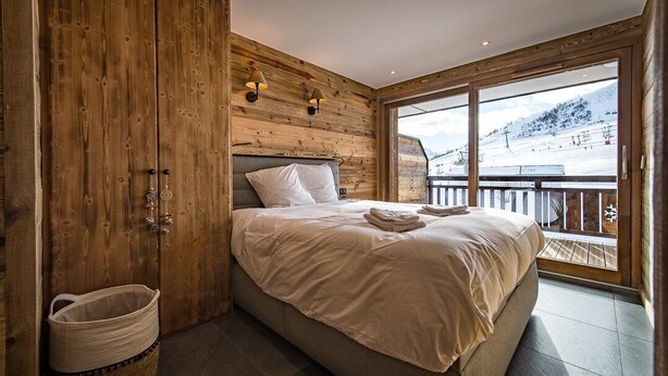 Ein Schlafzimmer mit einem Holz-Kopfteil, einem großen Bett mit weißer Bettwäsche, einem Holzschrank und Blick auf eine verschneite Landschaft durch ein großes Fenster.