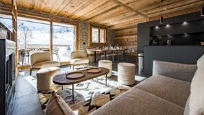 Ein gemütliches Blockhaus-Interieur mit Holzbalkendecke, einem Essbereich mit Tisch und Stühlen, einer Couch und einer Küchenzeile mit Arbeitsplatte und Pendelleuchten.