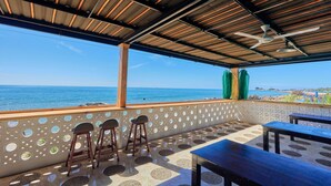 Un espace extérieur aménagé avec des sièges et offrant une vue sur la mer, caractérisé par un plafond en bois, un sol à motifs, ainsi qu’une table et des chaises.