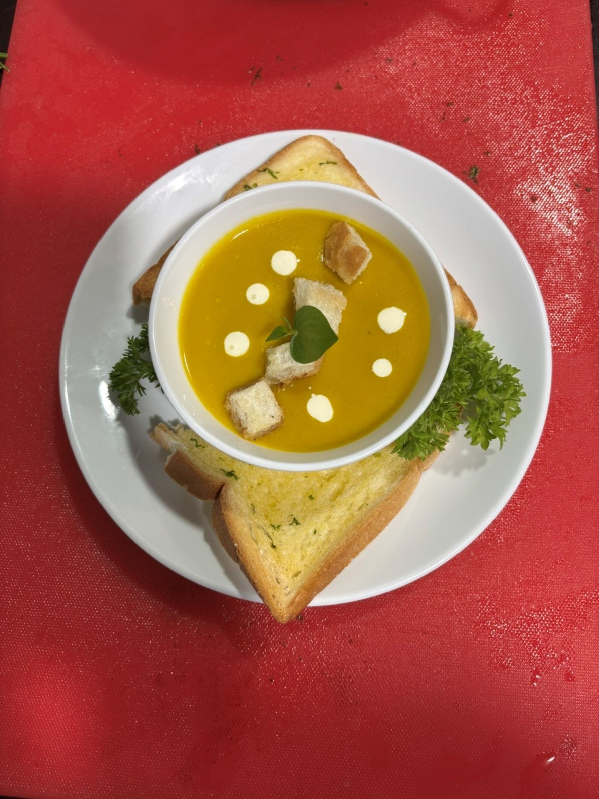 Una ciotola bianca con zuppa, crostini e cucchiaiate di panna su un tavolo con tovaglia rossa.
