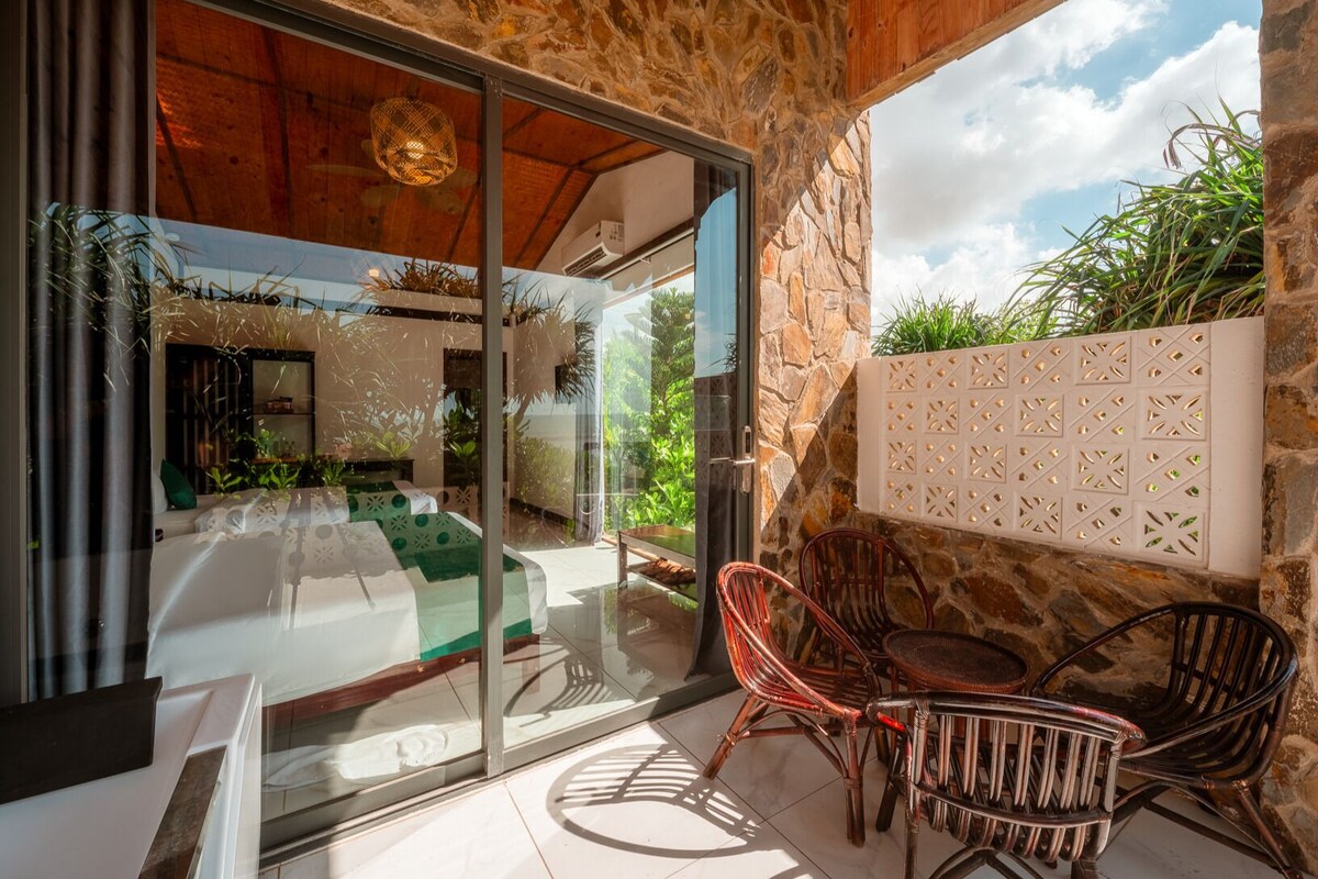 Een modern terras buiten, met een glazen deur naar een kamer, stenen muren en een zithoek met stoelen en een tafel.