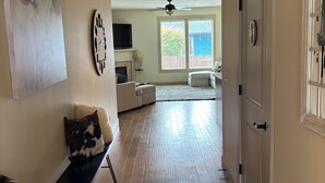 A spacious living room with hardwood flooring, a fireplace, a large window with blinds, a ceiling fan, and a door with a decorative handle.
