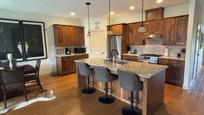 A modern kitchen with wooden cabinets, a granite island with bar stools, stainless steel appliances, and pendant lighting.