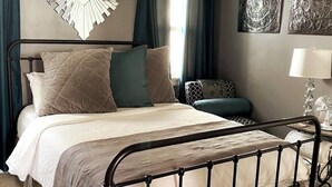 Una cama bien hecha con estructura metálica, un espejo decorativo encima y una lámpara en la mesita de noche.