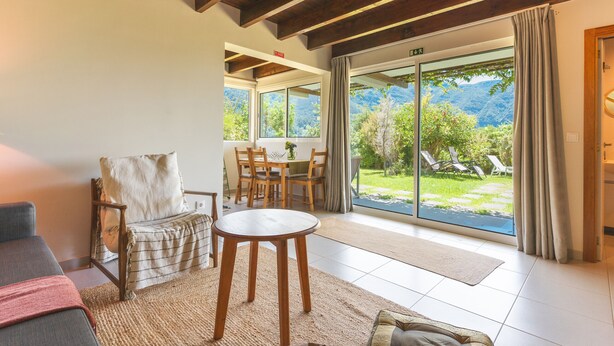 A room with a wooden ceiling, a sofa, a table, and a view of a garden through a glass door.