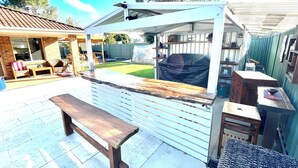 Un espace extérieur aménagé avec une terrasse, un banc en bois, un coin salon abrité et un barbecue.