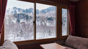 A room with a large window offering a view of a snowy forest. There is a wooden table and a cushioned bench. - Ishinoyu Lodge (Yamanouchi)