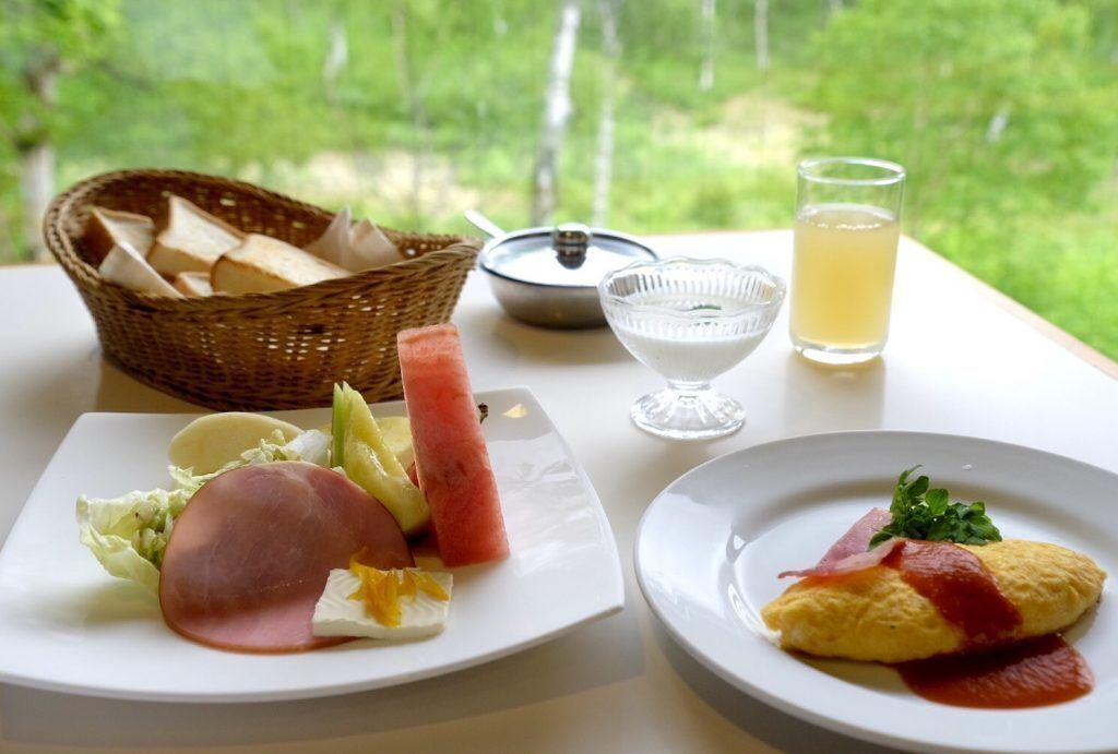 A table with breakfast items including ham, fruits, and an omelette, with a view of greenery in the background.