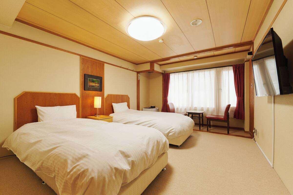 Una camera d'albergo con due letti, una scrivania, una sedia, una televisione e una finestra con tende.