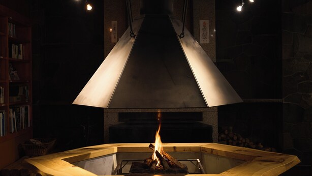 A wood-burning stove with a large, conical chimney, surrounded by bookshelves and a stone wall. - Ishinoyu Lodge (Yamanouchi)
