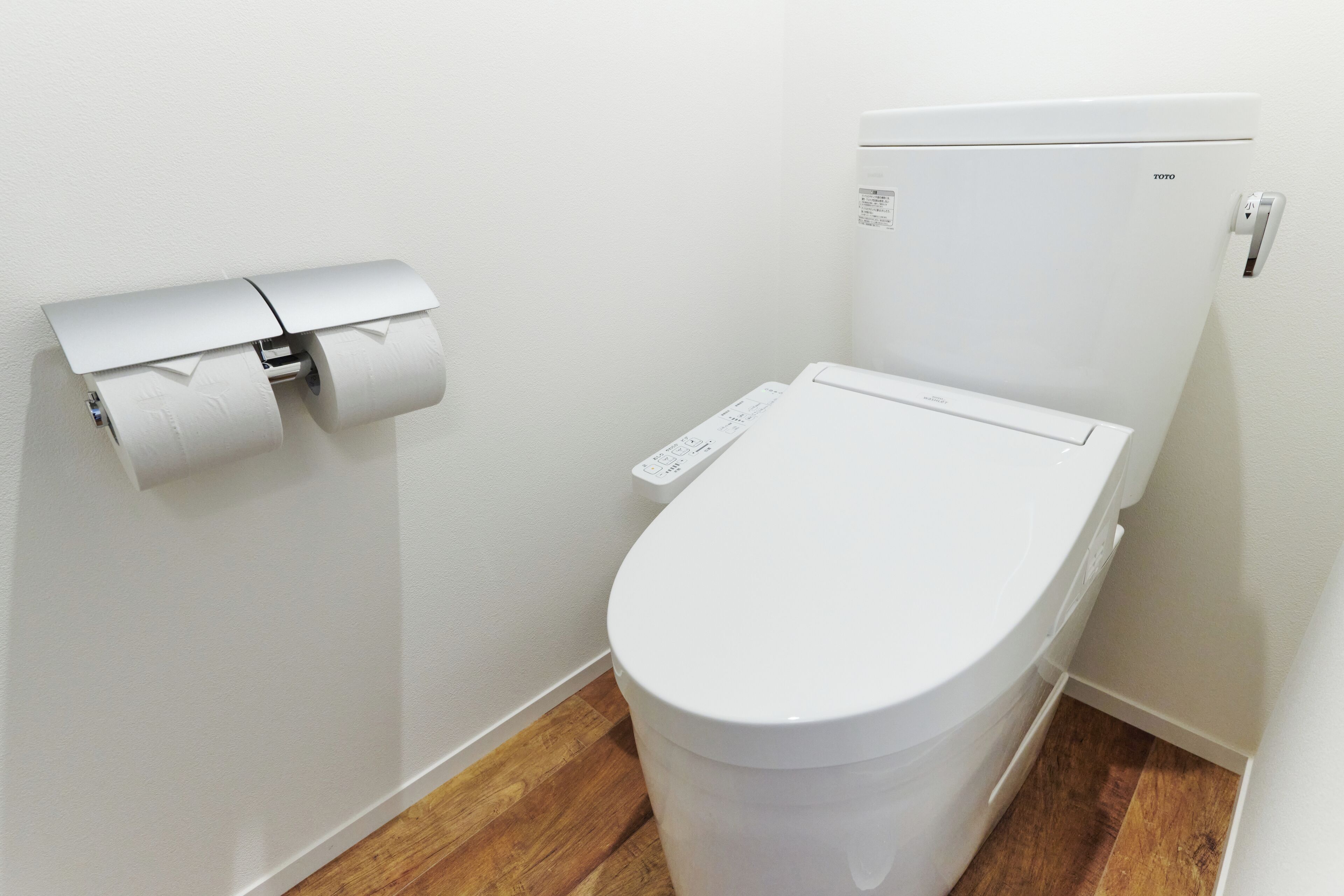 Sanitário branco com painel de controle, rolo de papel higiênico na parede e piso de madeira.